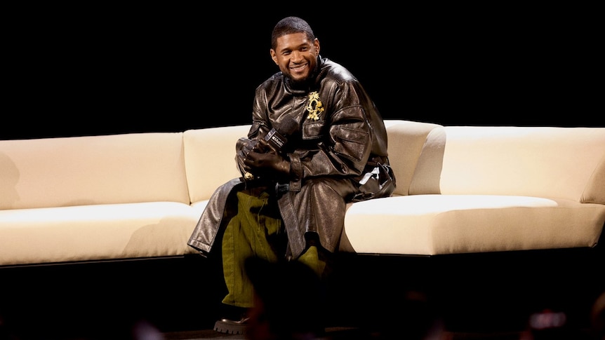 Usher sitting on a couch dressed in all black, smiling and leaning forward slightly