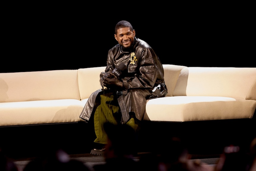 Usher sitting on a couch dressed in all black, smiling and leaning forward slightly