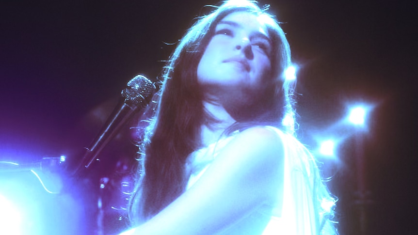 Weyes Blood plays piano looking upward in a shot with vaseline lens and piercing blue light