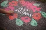 A wreath with red flowers and green leaves drawn in chalk on a footpath. 'Lest we forget' is in white chalk in the middle.
