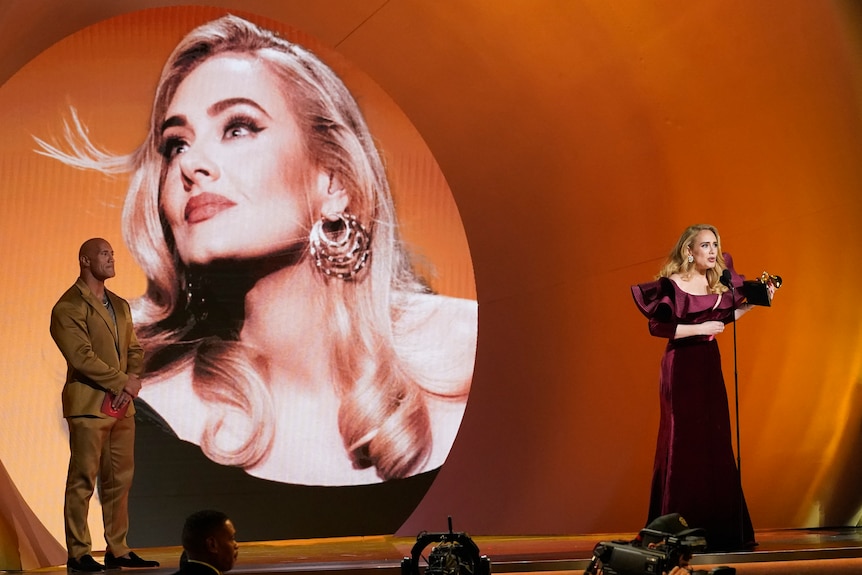 Adele holds a trophy and speaks into a microphone while a screen behind her displays a picture of her head. The Rock looks on.