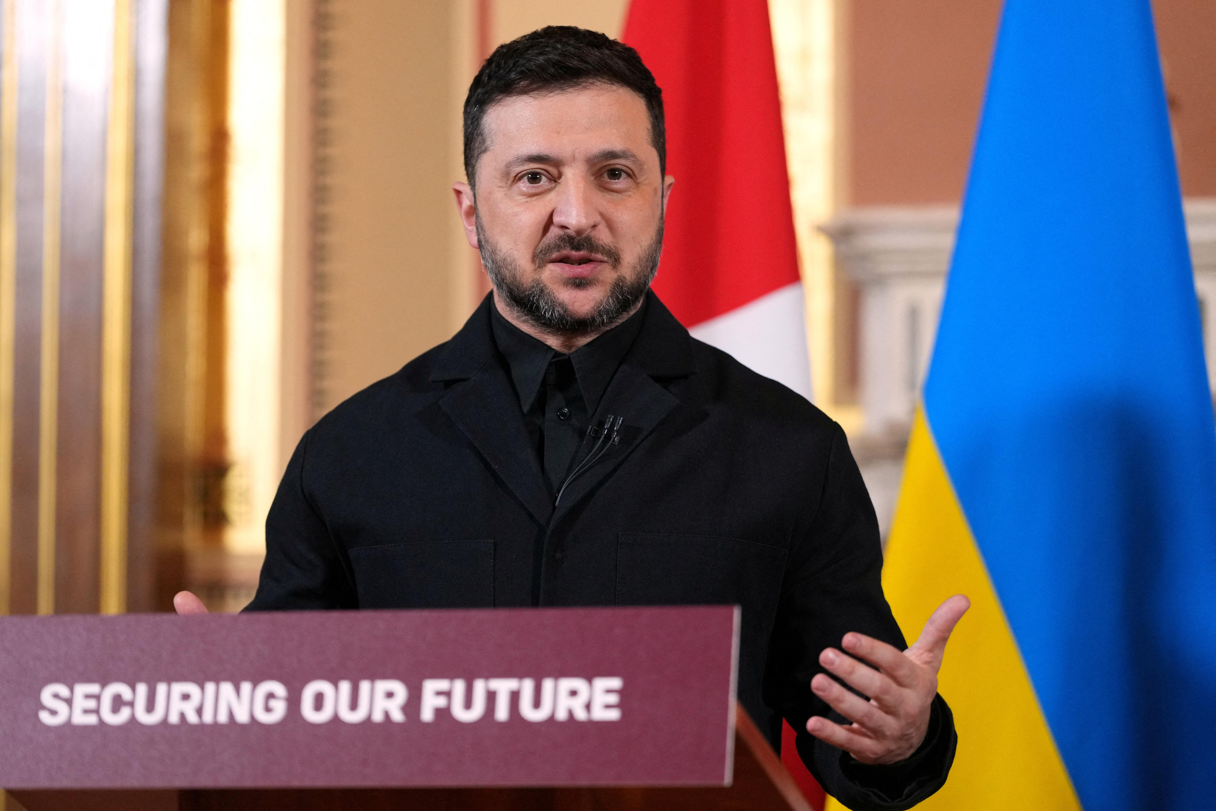 A man in a black top with a dark beard talks in front of brightly coloured flags while standing at a lectern.