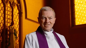 Father Kevin Dillon stands smiling in a church, wearing his priest's robes.