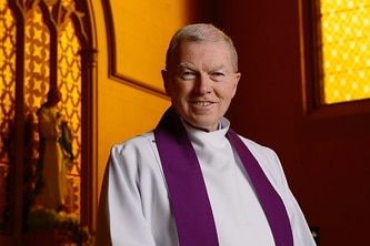 Father Kevin Dillon stands smiling in a church, wearing his priest's robes.