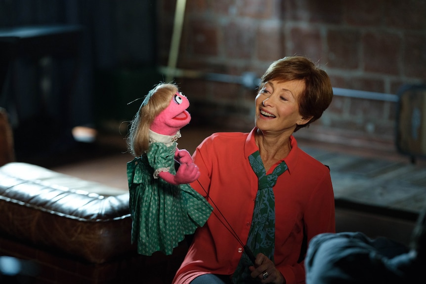White woman with short brown hair wears read shirt and holds pink-skinned puppet with blonde hair and green dress.