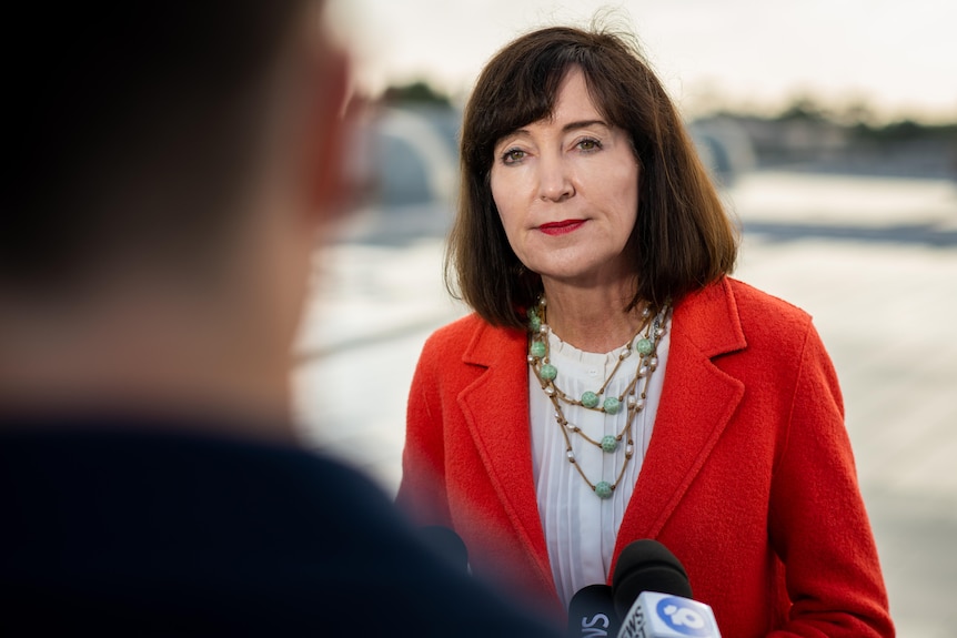 A woman with brown hair, wearing a red blazer with a neutral expression