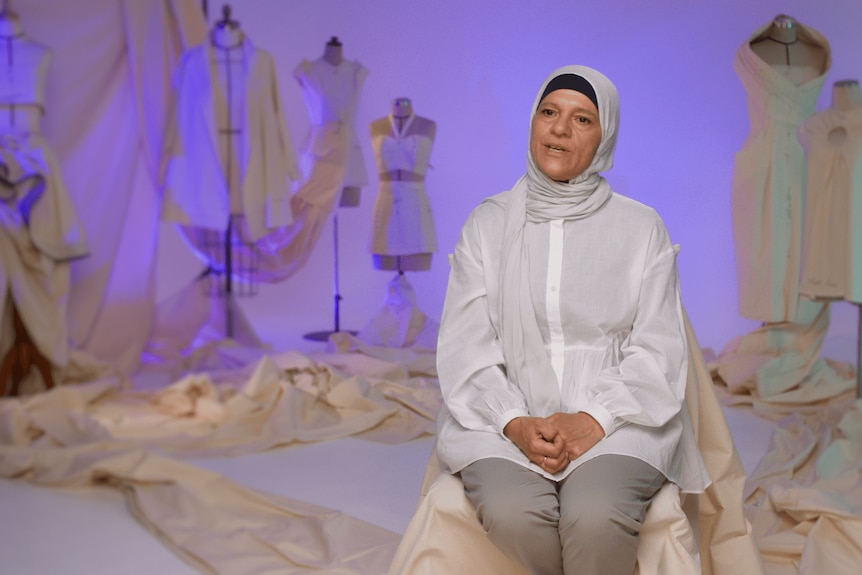 Aheda, wearing a white blouse and a grey hijab sits on a TV set with fashion bodice models and fabric in the background