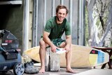 Dave Porter sits on the deck of his surfboard shaping shipping container, sanding a fin.