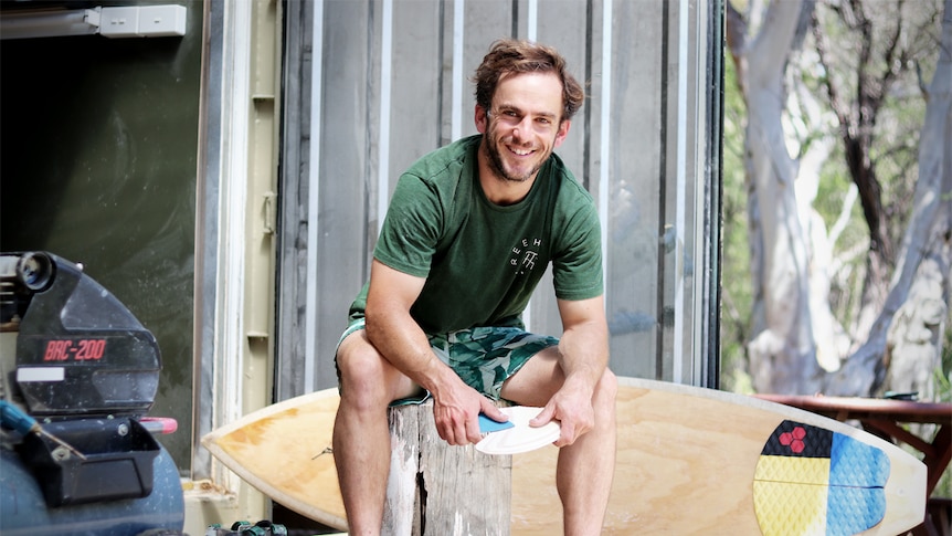 Dave Porter sits on the deck of his surfboard shaping shipping container, sanding a fin.