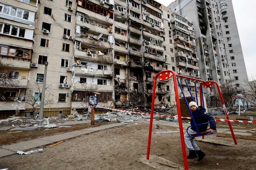 A child sits on a swing in front of a damaged residential building.