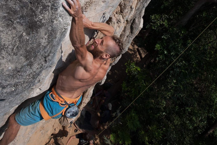 A muscular man without a shirt climbs the exterior of a large rock