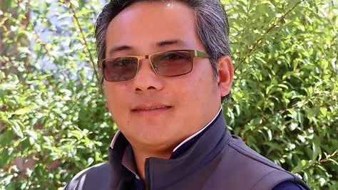 A man with short, silvery black hair stands outside in a winter vest.