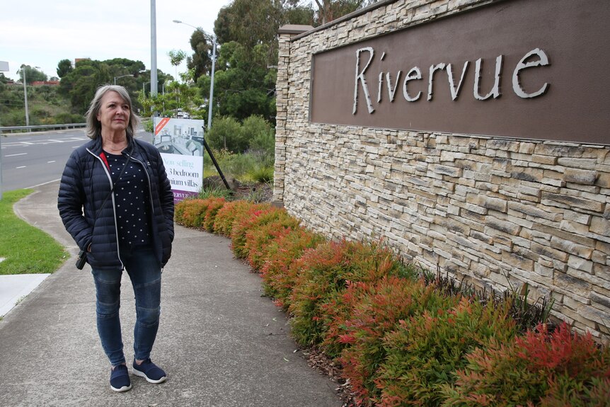 A woman wearing dark clothing stands next to a wall bearing a sign saying 'Rivervue'.