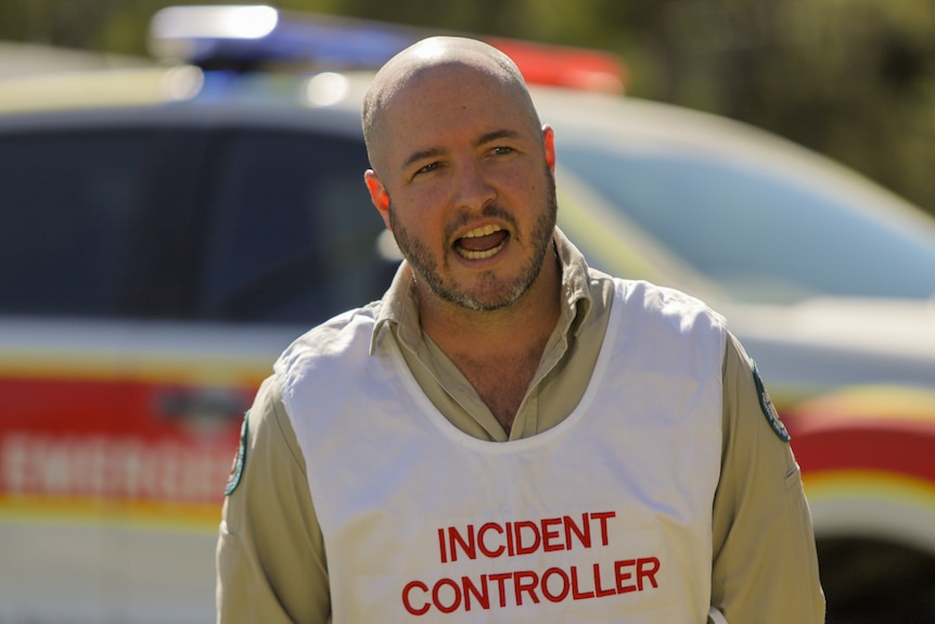 A bald man in a white vest with red writing that says: INCIDENT CONTROL speaking. 