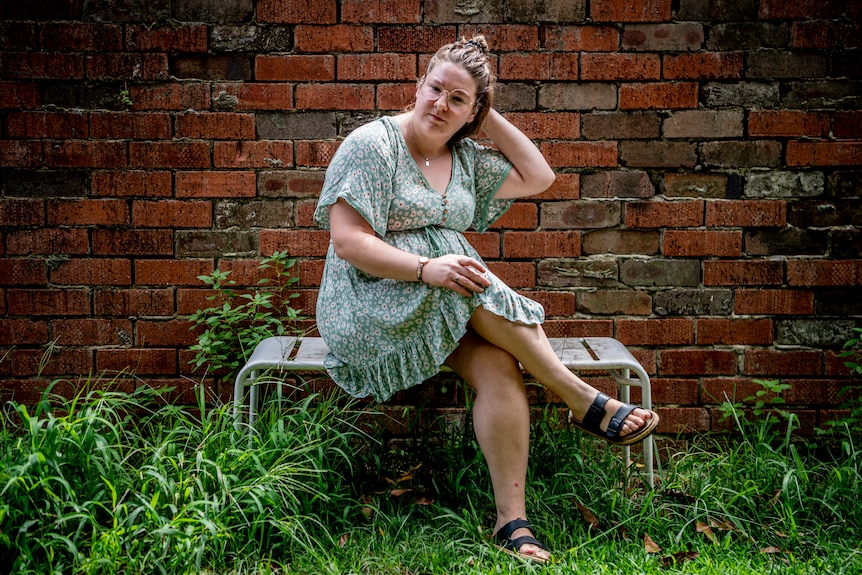 A young white woman with glasses in a green dress, sitting in front of a brick wall