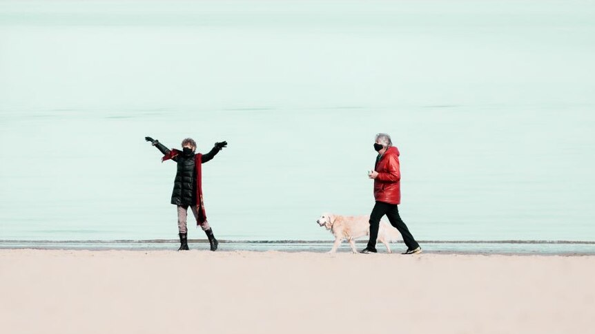 Two people in puffer jackets and masks stand on a beach with a golden retriever. One woman stretchers her arms in the air.