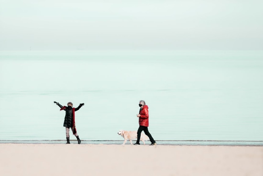 Two people in puffer jackets and masks stand on a beach with a golden retriever. One woman stretchers her arms in the air.