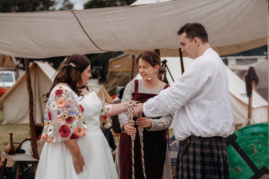 A bride and groom hold hands while a celebrant ties their hands together using a braid.
