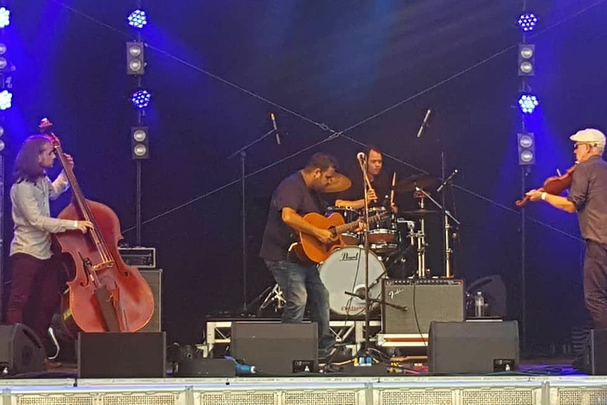 Stand up bass player, guitarist, drummer and violin player on stage with blue background lighting