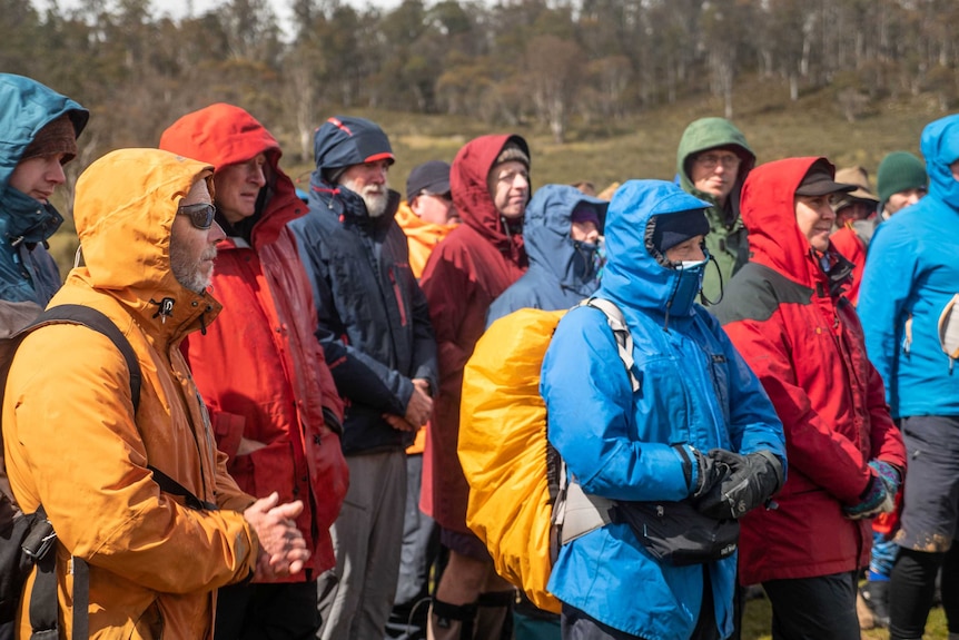 A group of protestors all wearing colourful rain jackets, orange, red blue and green, trees on a hill in the backdrop.