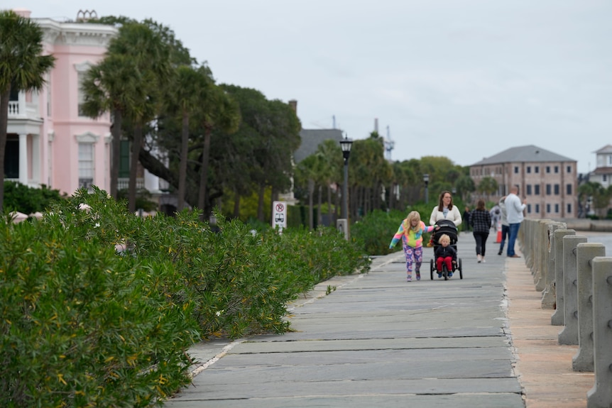 Families walk along a wooden boardwalk beside oceanfront homes with a grey sky above.