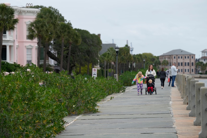 Familiile se plimbă de-a lungul unei promenade lângă case cu vedere la ocean, cu un cer gri deasupra.