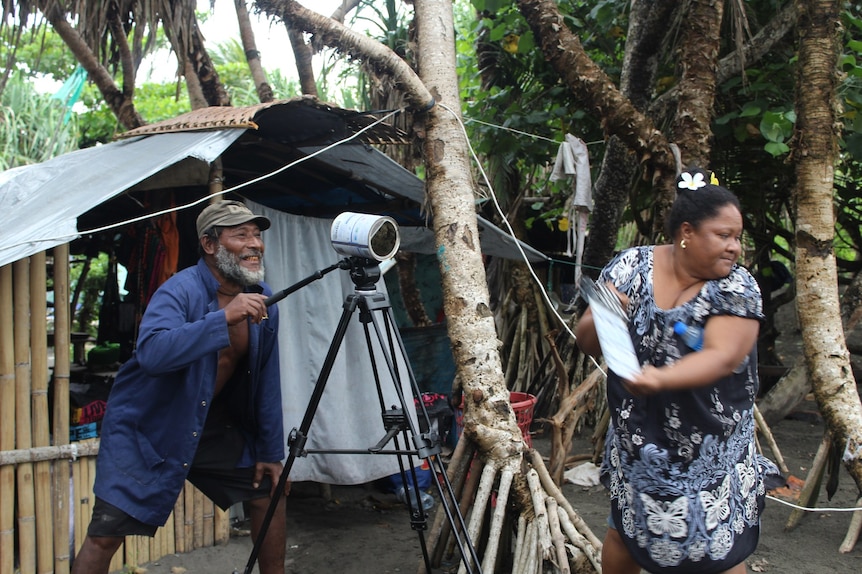 Man holds camera tripod mouted with tin behind a woman holding clapper board in PNG village. 