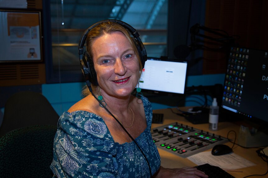 Blonde woman in a blue dress behind a mixing desk smiles at camera 