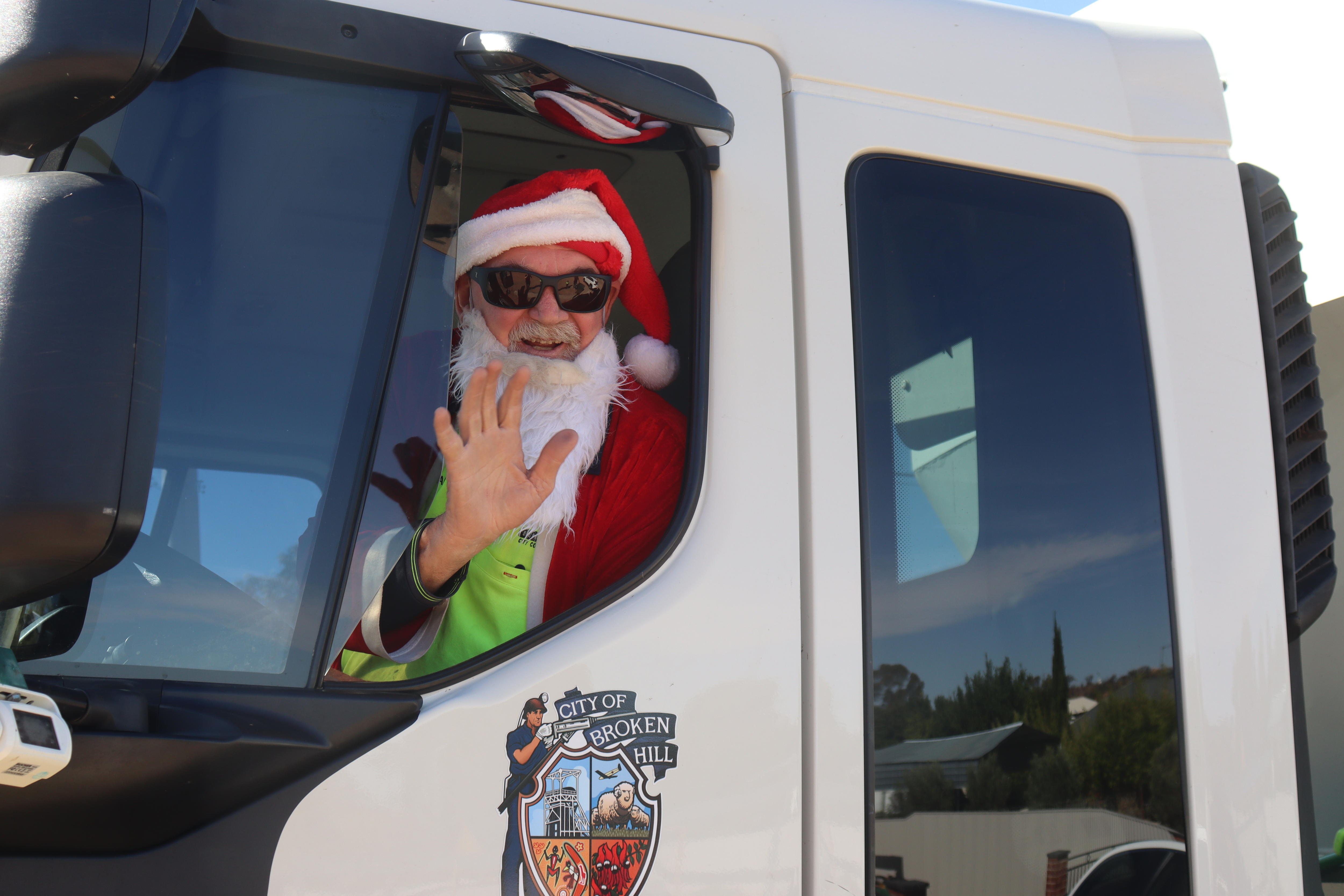 A man wearing a fake Santa beard and jacket waves from a garbage truck window.
