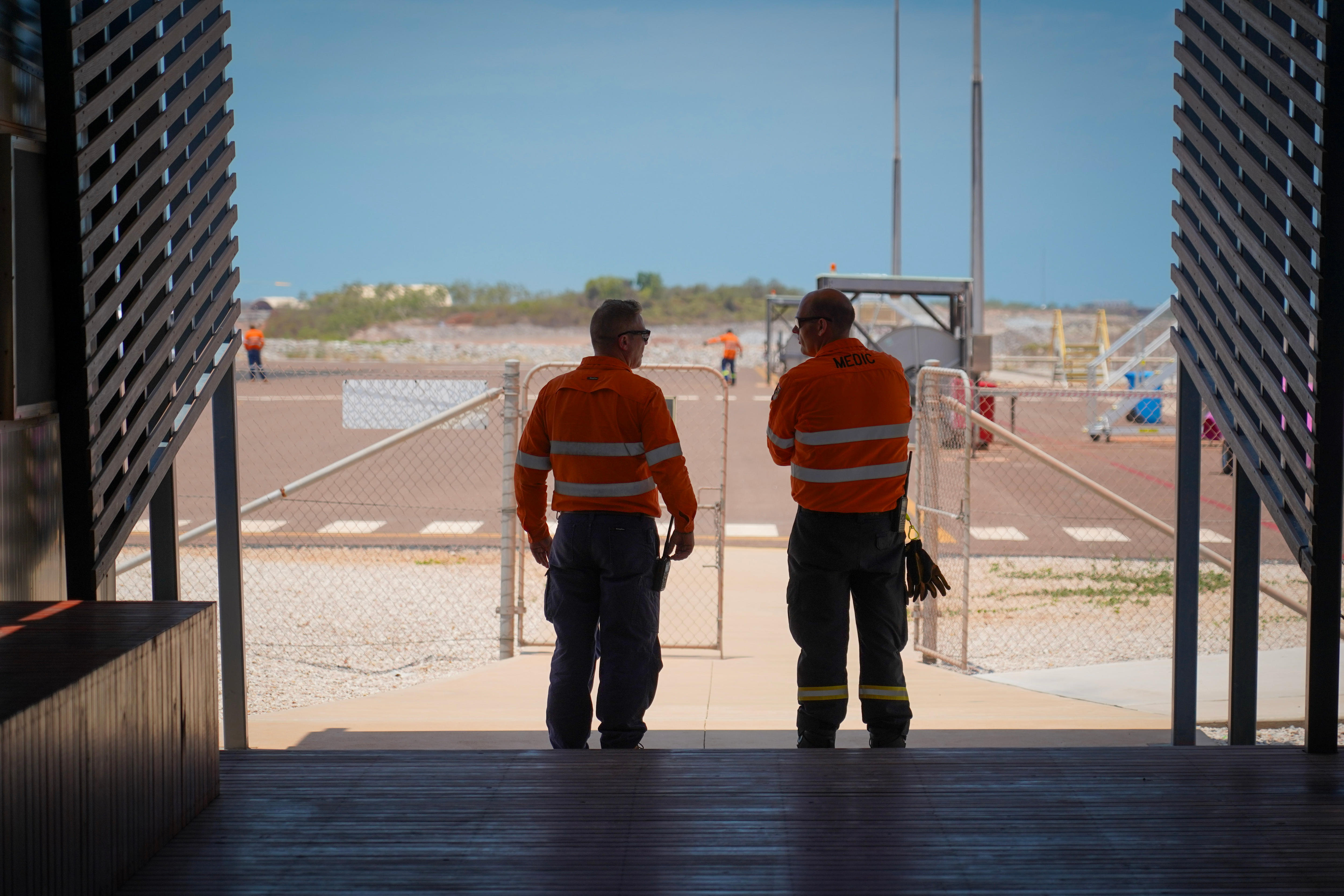 workers in hi vis stand in the shade of a shed overlooking the airstrip