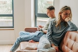 A couple sit next to each other on the couch. The man scrolls on his phone as the woman looks away, upset.