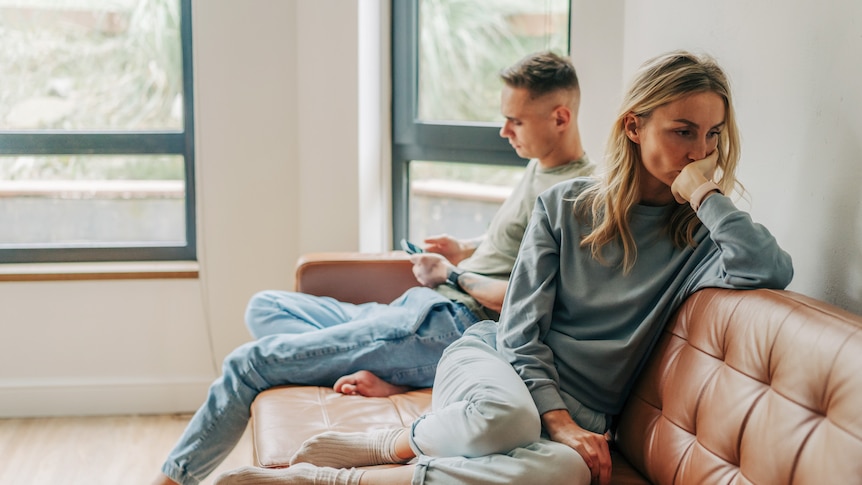 A couple sit next to each other on the couch. The man scrolls on his phone as the woman looks away, upset.