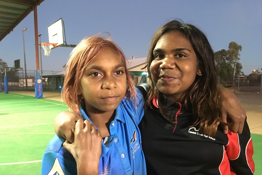 The two girls have their arms over each others shoulders, and are pictured by a basketball court