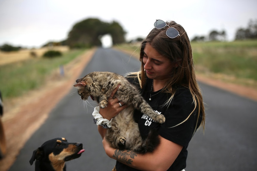 A woman stands on a road holding her cat while her dog looks on.