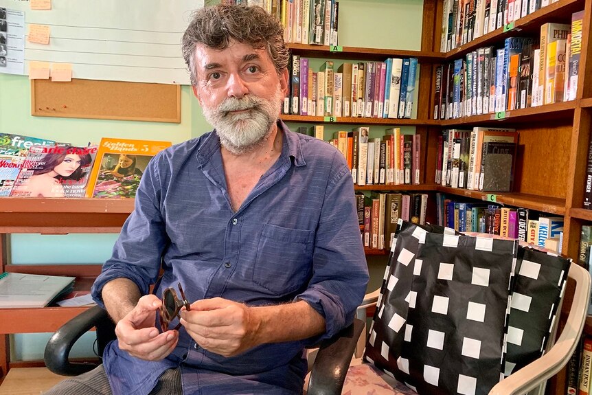 A man sits in front of bookcases and holds sunglasses as he poses for a photograph.