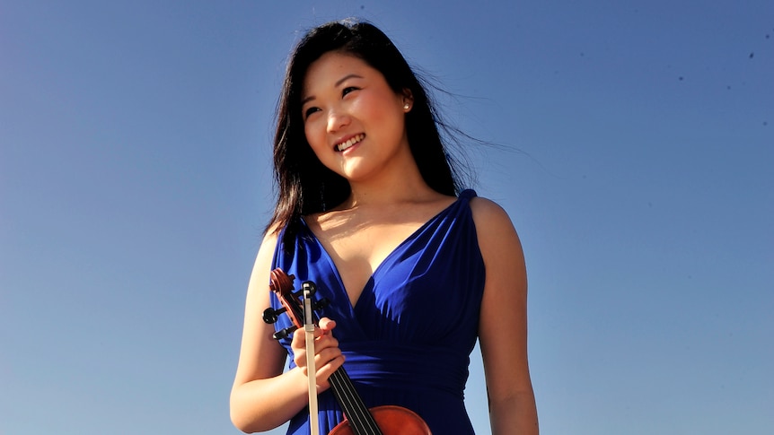 Violinist Emily Sun standing in a field wearing a blue dress holding her violin against a blue sky