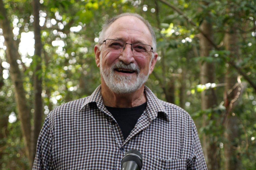 A man in his 70s smiling in front of a forest, short grey beard and glasses.