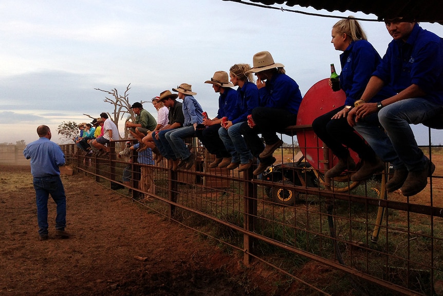 A man talks to a bunch of people who are sitting on a cattle yard rail