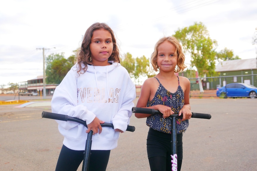 Two young girls standing on scooters smiling. 