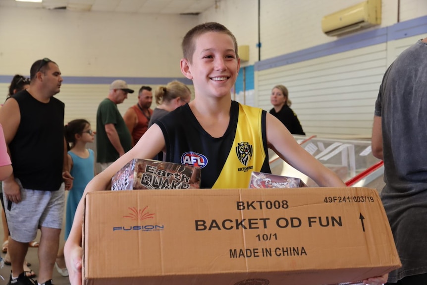 A young teen boy carrying a brown cardboard box with fireworks in it, smiling.
