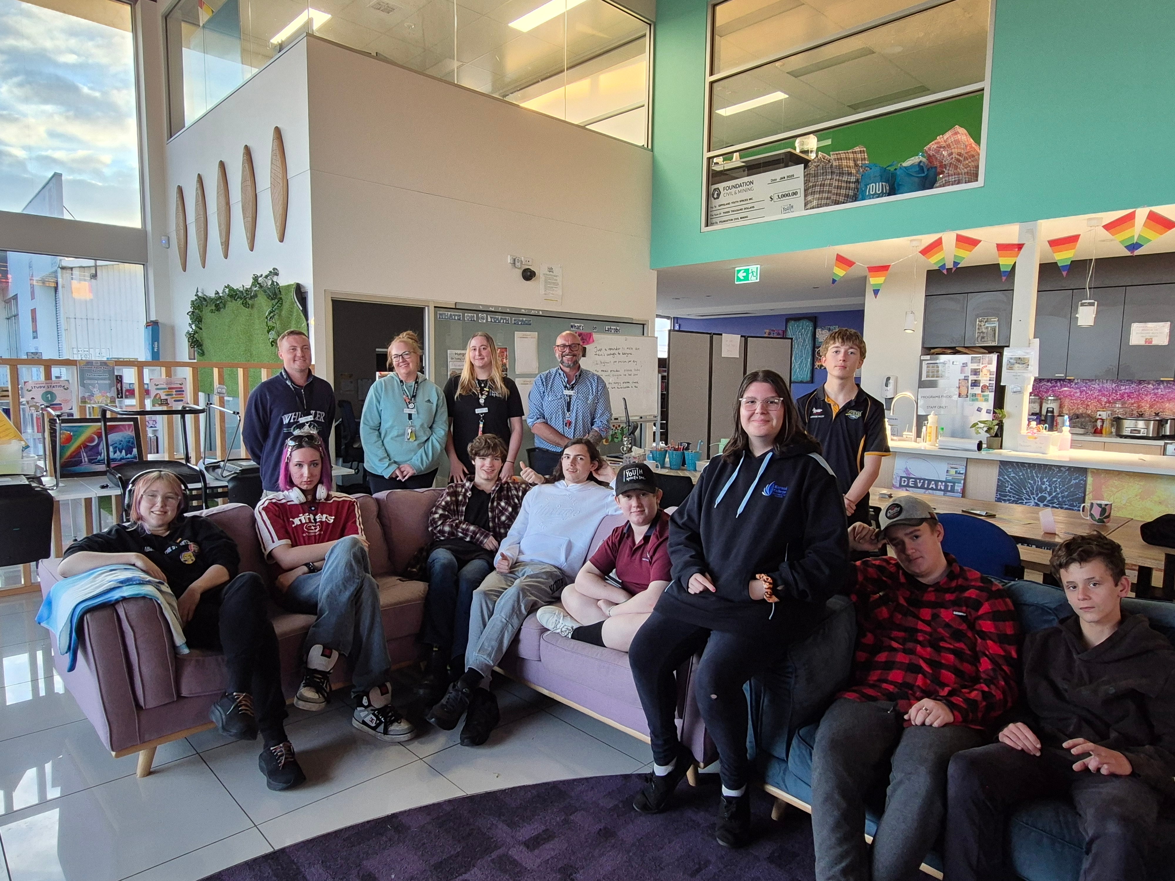 A group of young people in casual clothes sitting on a sofa in a big building, with adults standing behind them.
