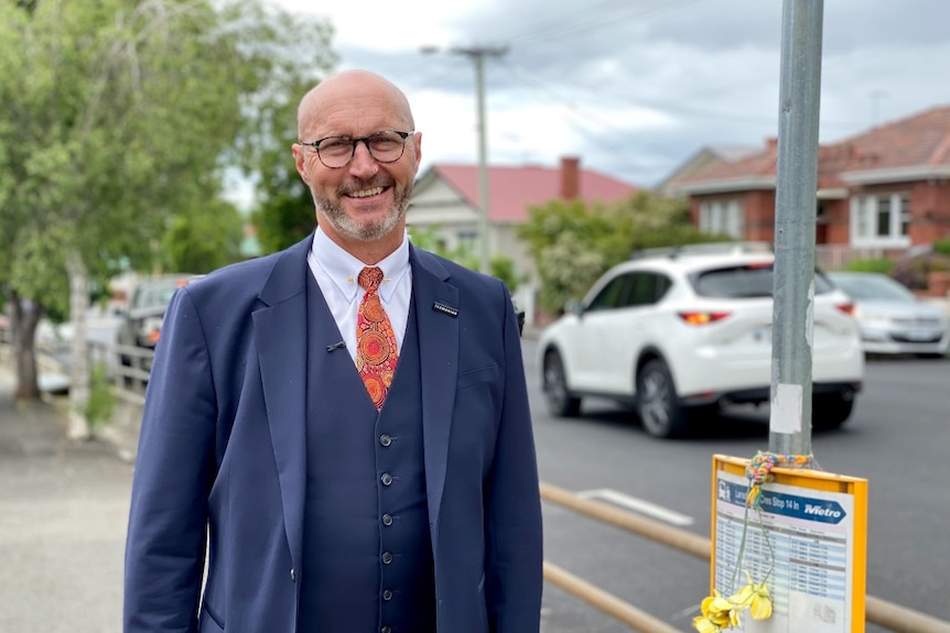 A balding man in a suit and glasses stands on a suburban street