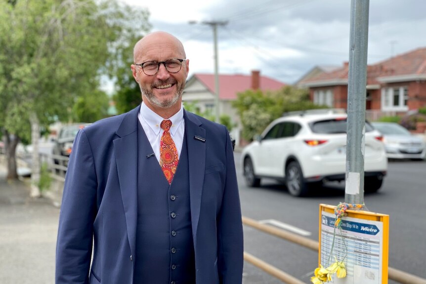 A balding man in a suit and glasses stands on a suburban street