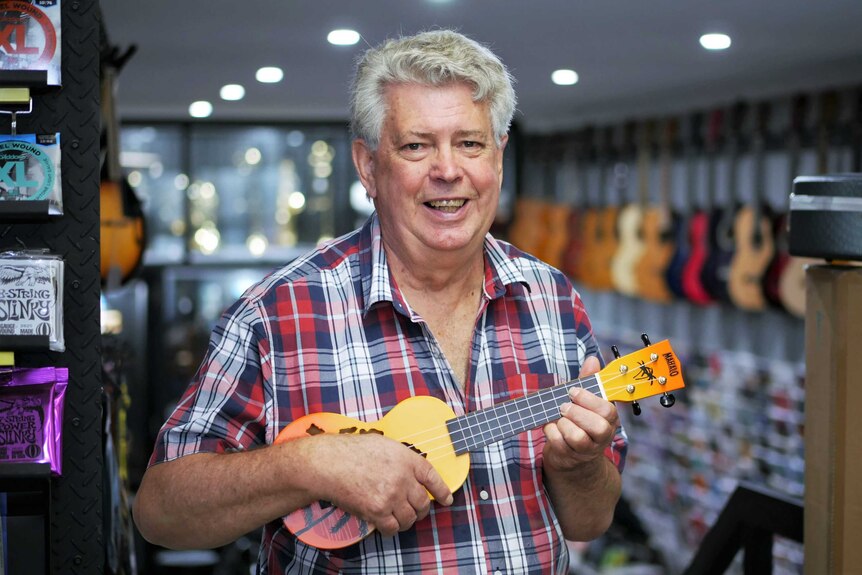 A man stands smiling while holding a brightly coloured ukulele in a music shop.