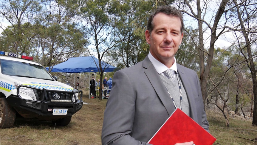 Detective Acting Inspector Craig Fox wearing a grey suit stands in front of a police vehicle in bushland.