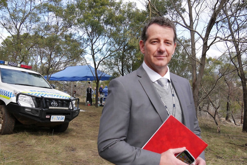 Detective Acting Inspector Craig Fox wearing a grey suit stands in front of a police vehicle in bushland.