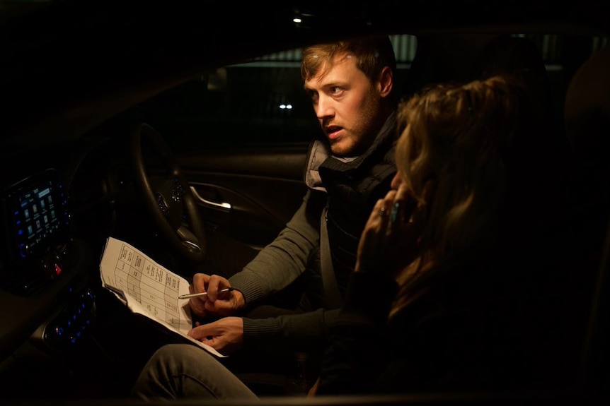 A dark photo of Anne-Marie and Chris sitting in a car at night. She takes a phone call. Their faces are illuminated by light.