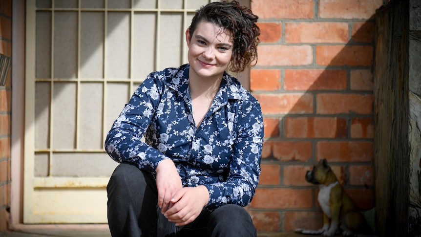 A woman wearing a patterned blue shirt and black jeans sits on the step outside a building.