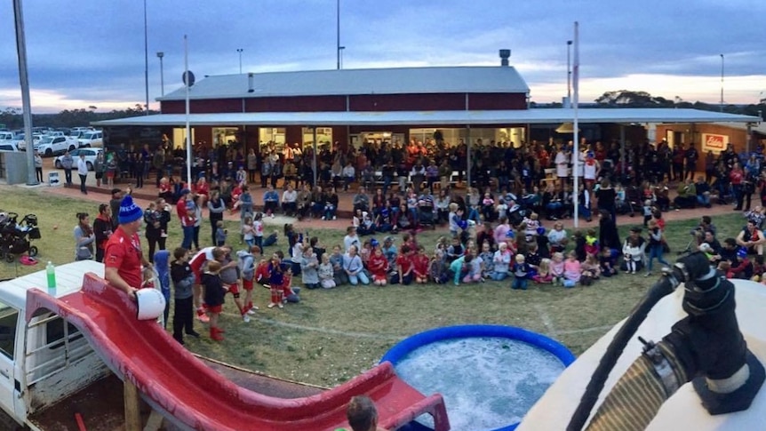 A large crowd sitting and standing around a ute and paddling pool full of water.