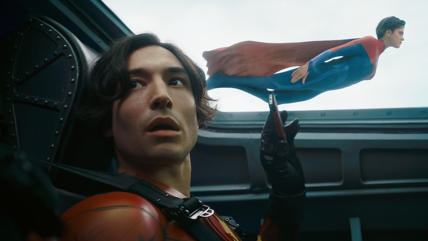 Ezra Miller, a dark-haired nonbinary person, sits in a jet. Outside, Gal Gadot, a brunette woman in superhero outfit, is flying.